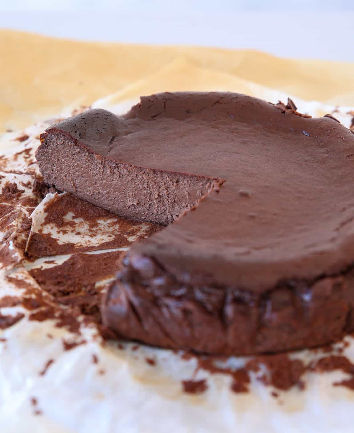 A rich, dense,  butrnt chocolate cheesecake with a slice removed sits on parchment paper, revealing its smooth, fudgy texture inside. Cocoa powder is scattered around the cake on the paper.