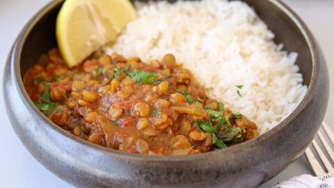 The Best Lentil Curry (Lentil Dahl) – Creamy & One-Pot A bowl of white rice served with lentil curry garnished with fresh herbs, accompanied by two lemon wedges. The dish is presented in a dark ceramic bowl.