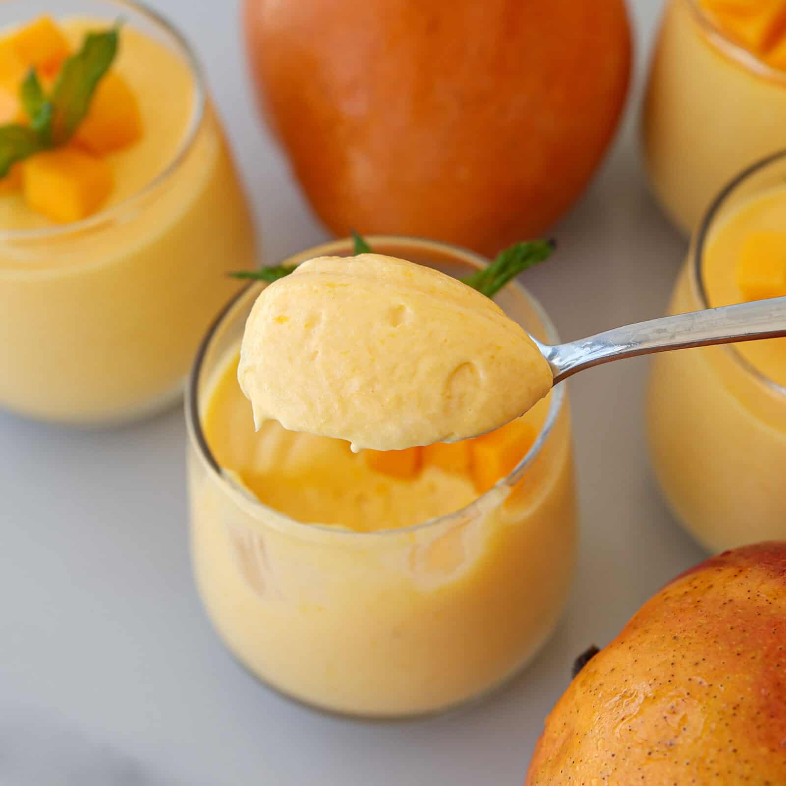 A spoonful of creamy mango mousse is held above a glass filled with more mousse; fresh mango chunks and mint leaves garnish the dessert, with whole mangoes visible nearby.
