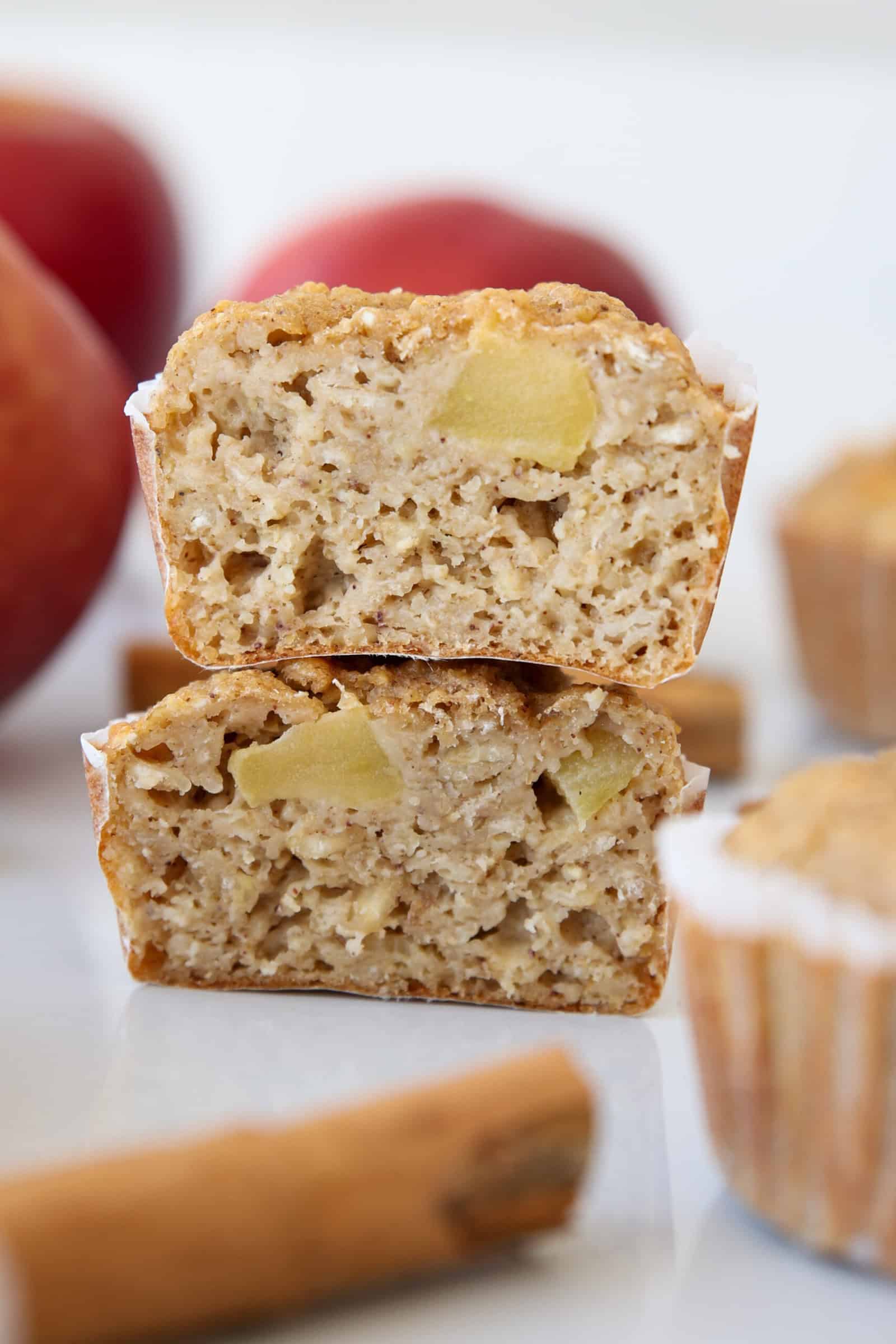 Two apple cinnamon muffins are stacked, cut in half to show a moist, dense interior with visible apple chunks. Cinnamon sticks and apples are blurred in the background.