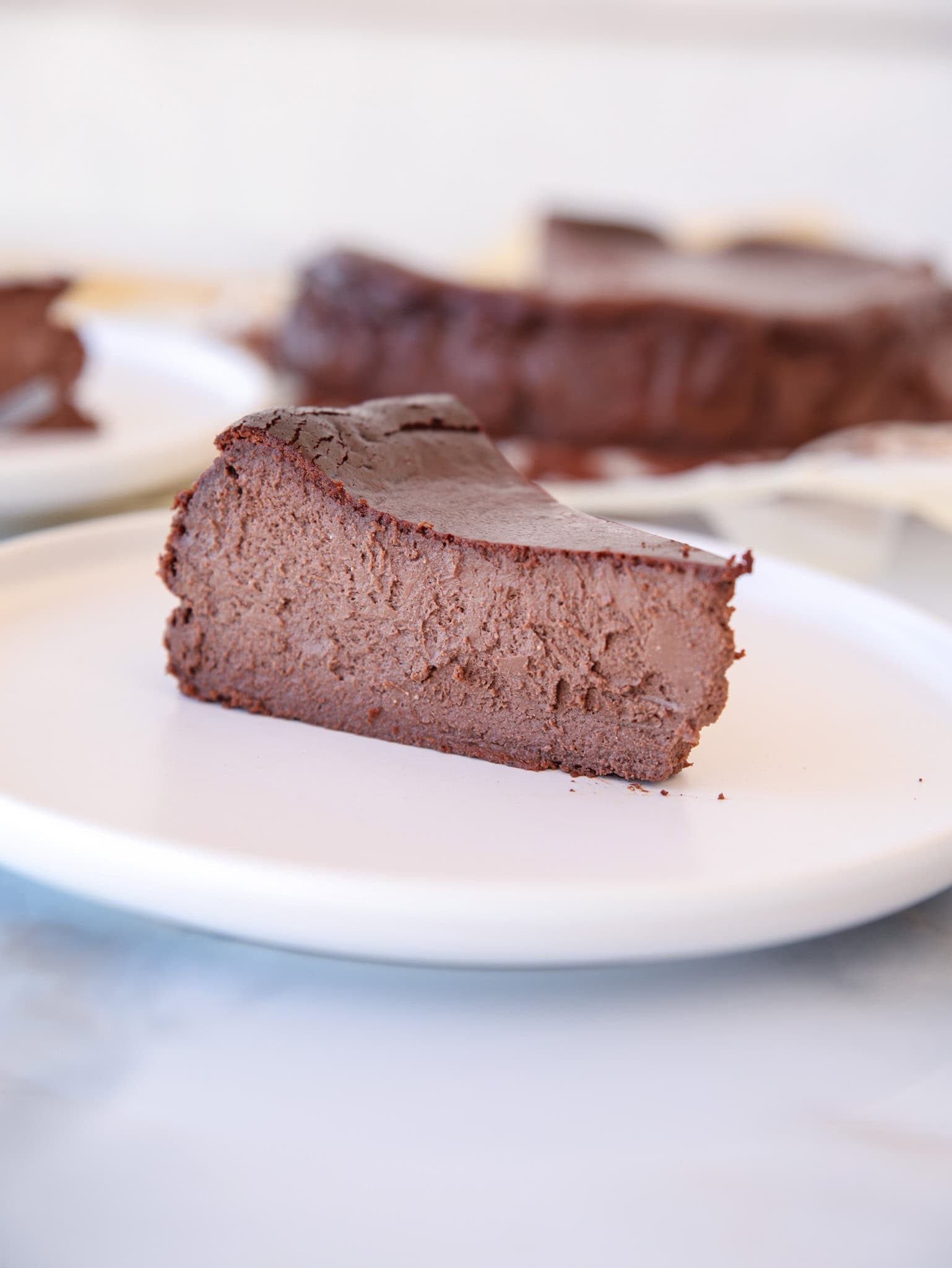 A thick slice of rich, creamy chocolate Basque cheesecake sits on a white plate, with the rest of the cheesecake visible in the blurred background.