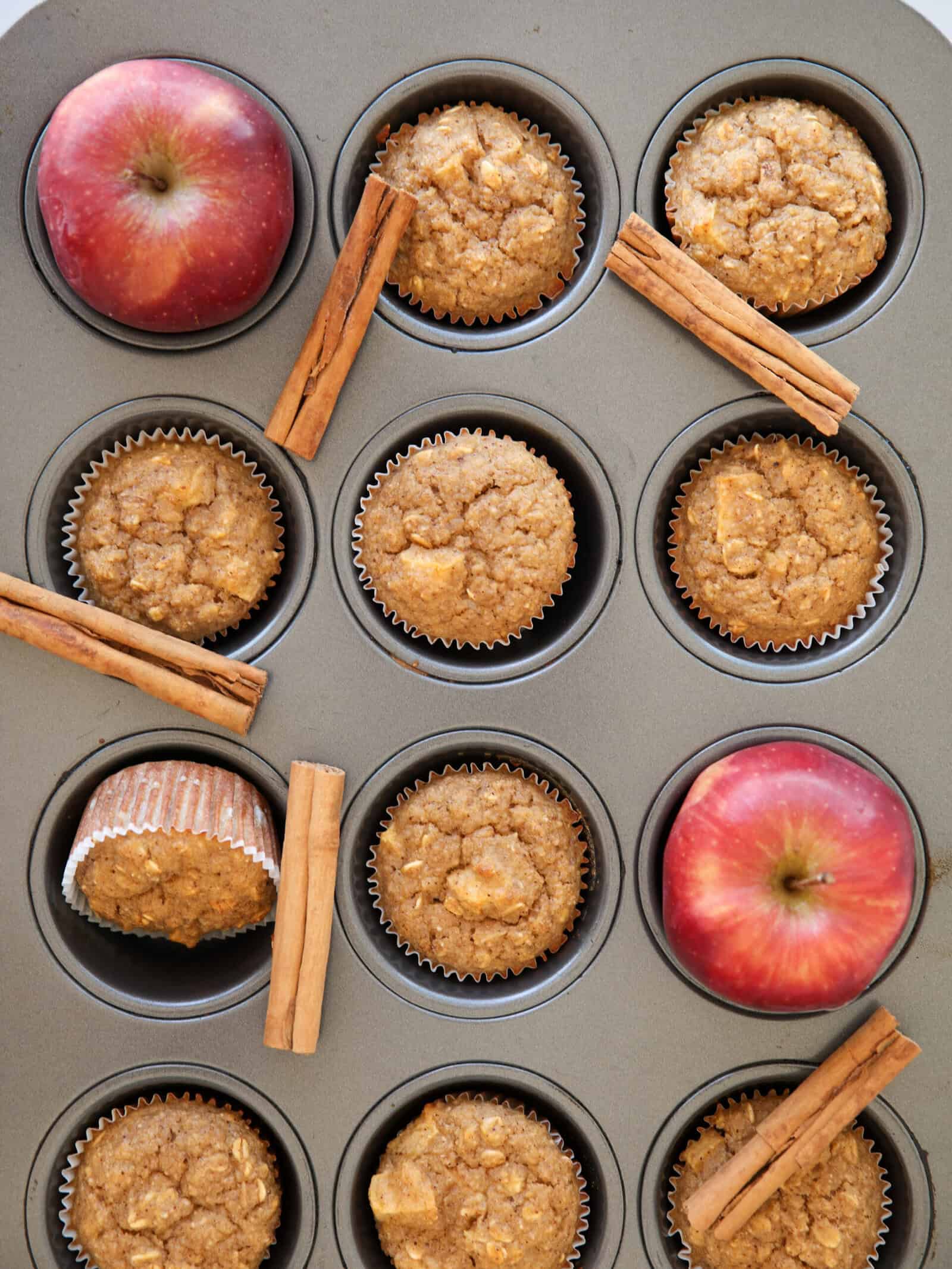 A muffin tin holds several baked muffins, whole red apples, and sticks of cinnamon. Some muffins are tilted, and the apples and cinnamon sticks are scattered among the muffins.