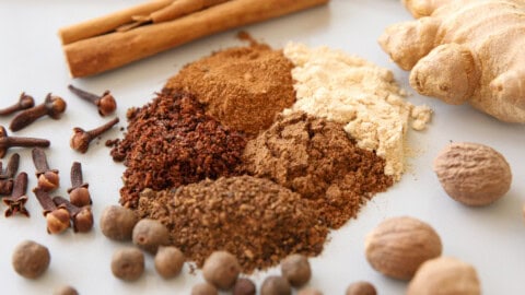 Easy Homemade Pumpkin Pie Spice Mix A close-up of assorted spices on a white surface, including ground spices in small piles, whole allspice, cloves, nutmeg, cinnamon sticks, and a piece of ginger root.