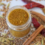 A glass jar filled with ground yellow curry spice blend, surrounded by whole dried spices including cinnamon sticks, cardamom pods, coriander seeds, dried red chilies, and cumin seeds.