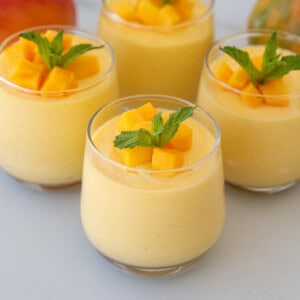 Four glasses of creamy mango mousse topped with fresh mango cubes and mint leaves, arranged on a light surface.