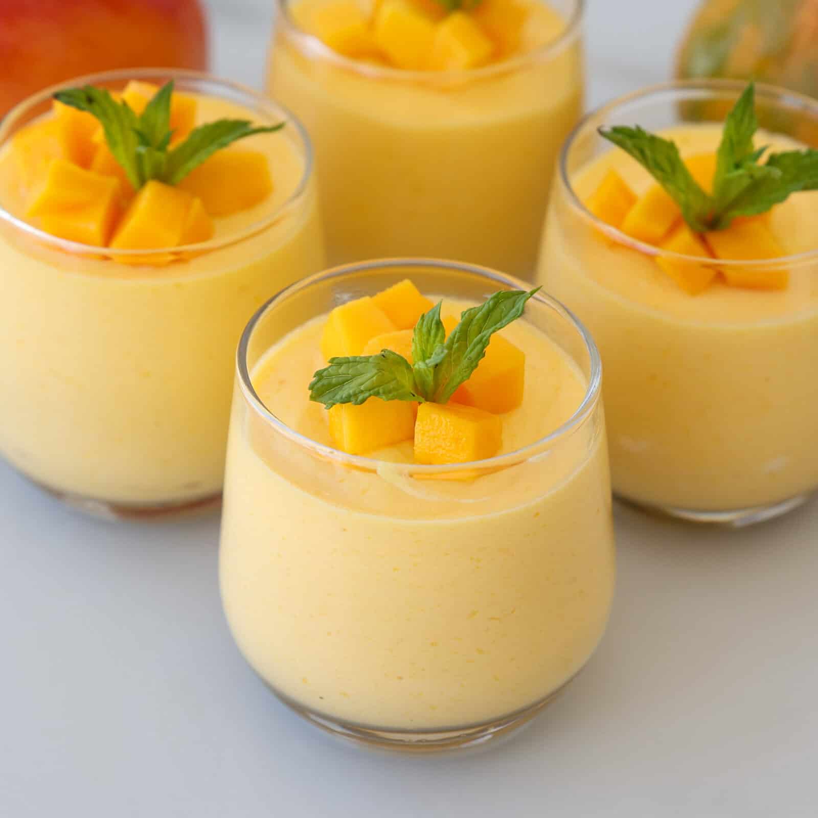 Four glasses of creamy mango mousse topped with fresh mango cubes and mint leaves, arranged on a light surface.