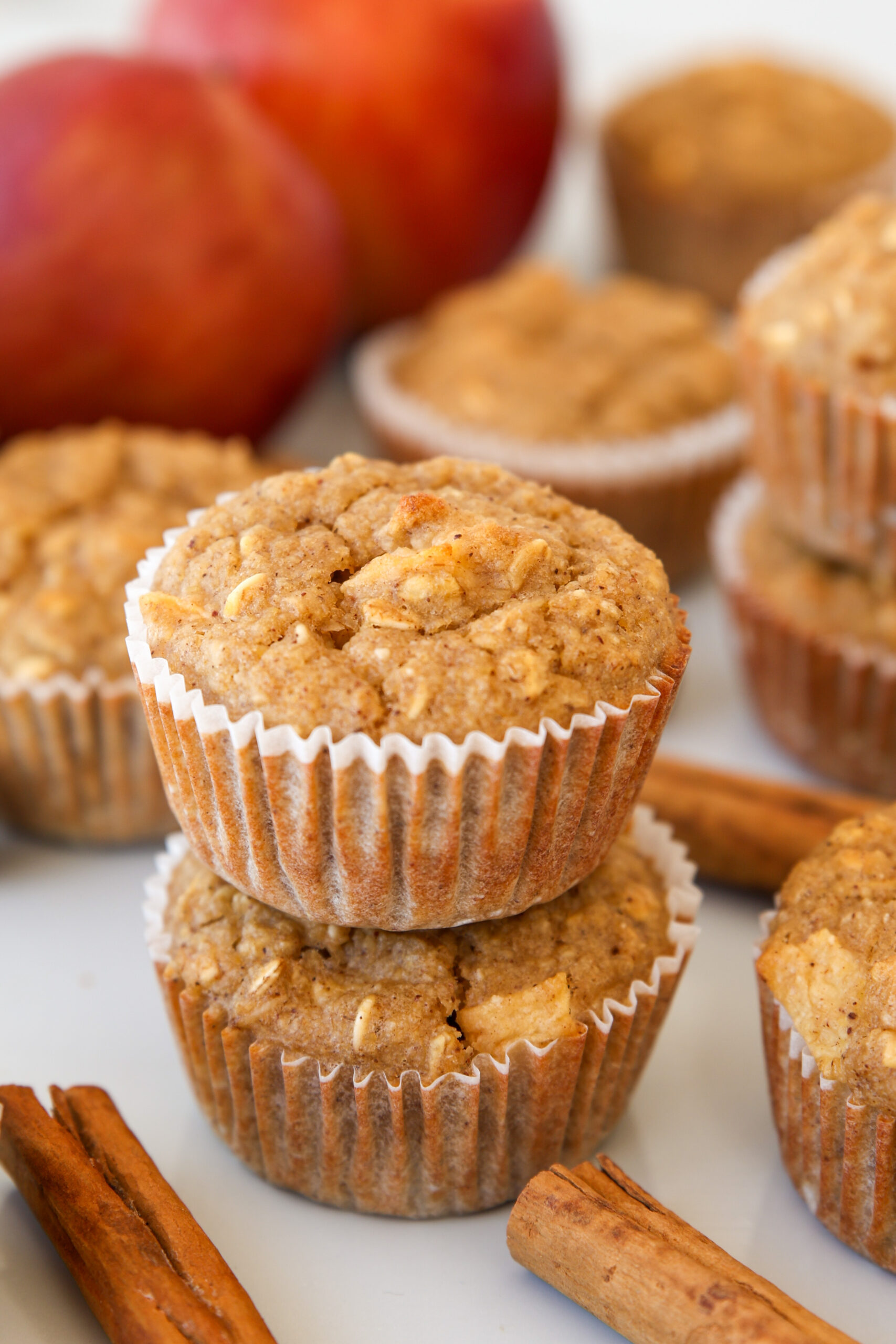 Two apple cinnamon muffins stacked on top of each other, surrounded by more muffins, cinnamon sticks, and whole apples in the background. The muffins are in white paper wrappers.