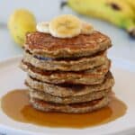 A stack of pancakes topped with banana slices and drizzled with syrup sits on a white plate, with whole bananas blurred in the background.