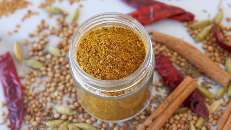 A glass jar filled with yellow curry powder is surrounded by whole spices, including dried red chilies, cinnamon sticks, green cardamom pods, coriander, and cumin seeds, scattered on a white surface.