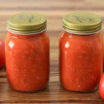 Two glass jars filled with homemade tomato sauce, sealed with gold lids, are placed on a wooden surface. Fresh, whole tomatoes surround the jars.