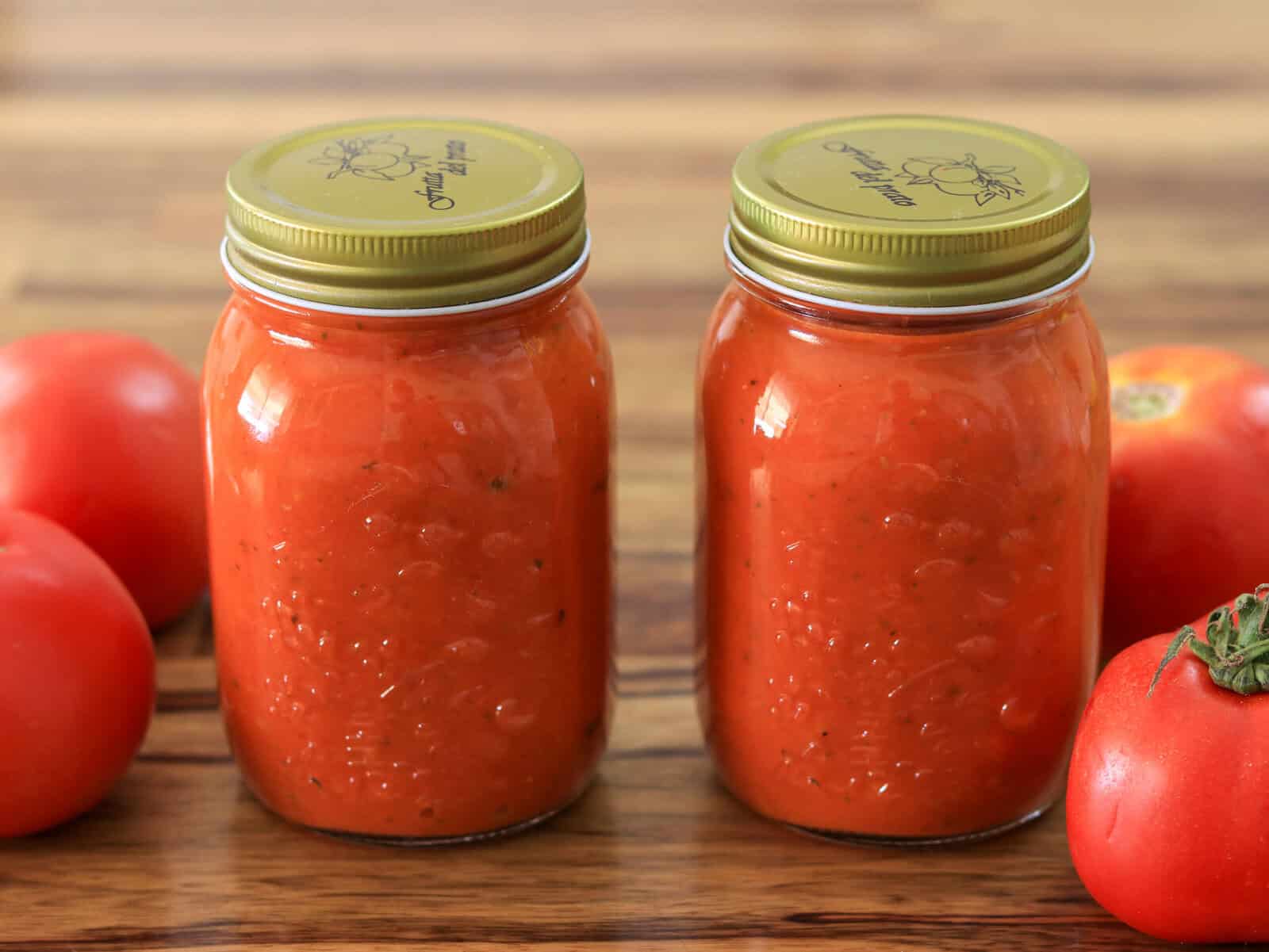 Two glass jars filled with homemade tomato sauce, sealed with gold lids, are placed on a wooden surface. Fresh, whole tomatoes surround the jars.
