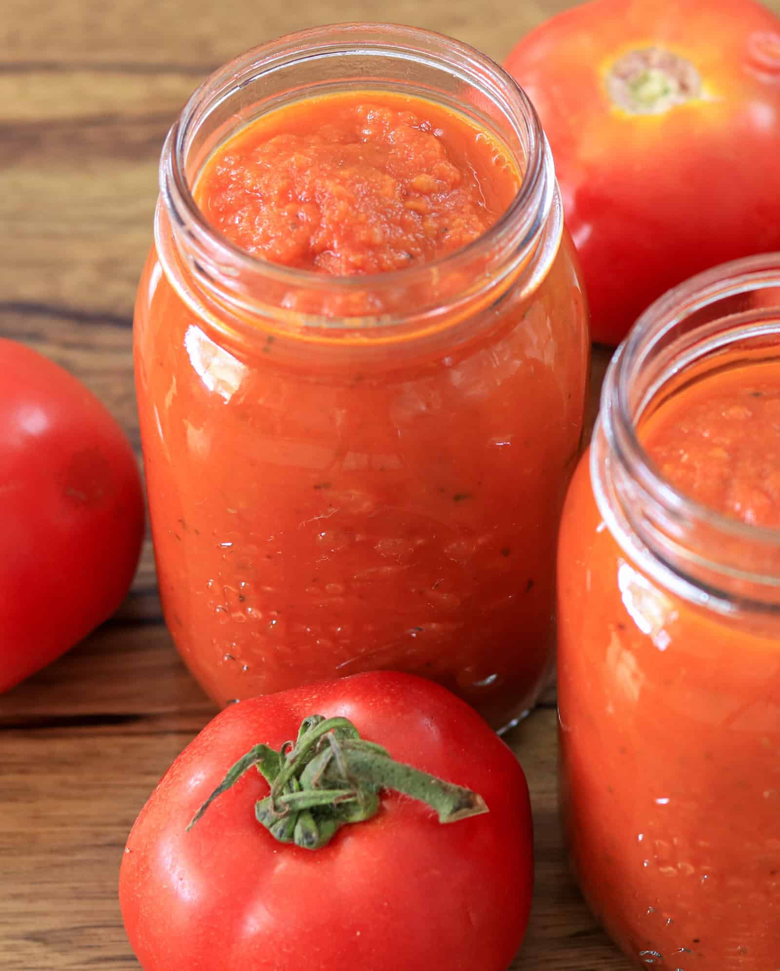 Two glass jars filled with homemade tomato sauce sit on a wooden surface, surrounded by fresh, whole tomatoes. The sauce is smooth and vibrant red-orange in color.