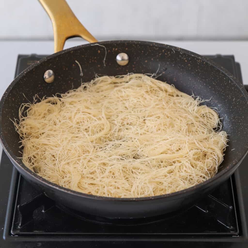 A nonstick frying pan with a layer of uncooked, thin vermicelli noodles sits on a stovetop, ready for cooking. The pan has a golden handle.