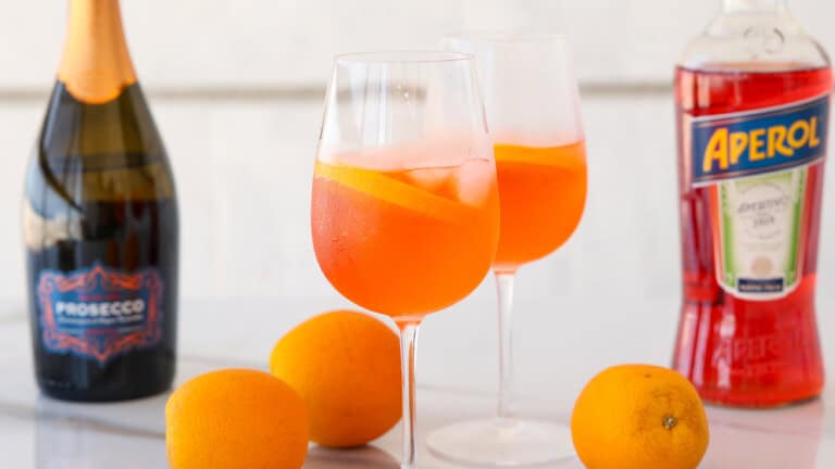 Two glasses of Aperol Spritz with ice and orange slices, surrounded by whole oranges, a bottle of Prosecco, and a bottle of Aperol on a white surface.