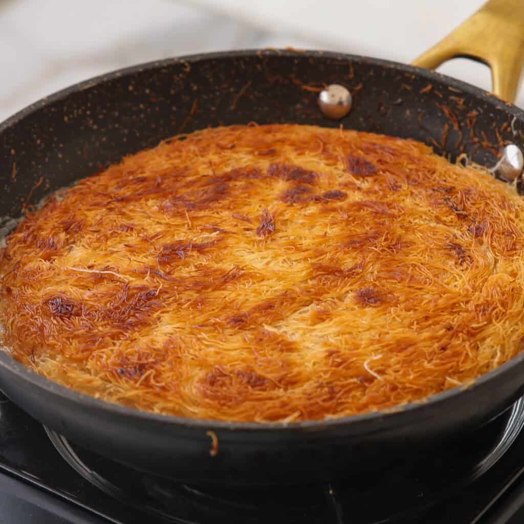 A golden-brown, crispy layer of shredded phyllo dough dessert cooks in a black frying pan with a yellow handle on a stovetop.