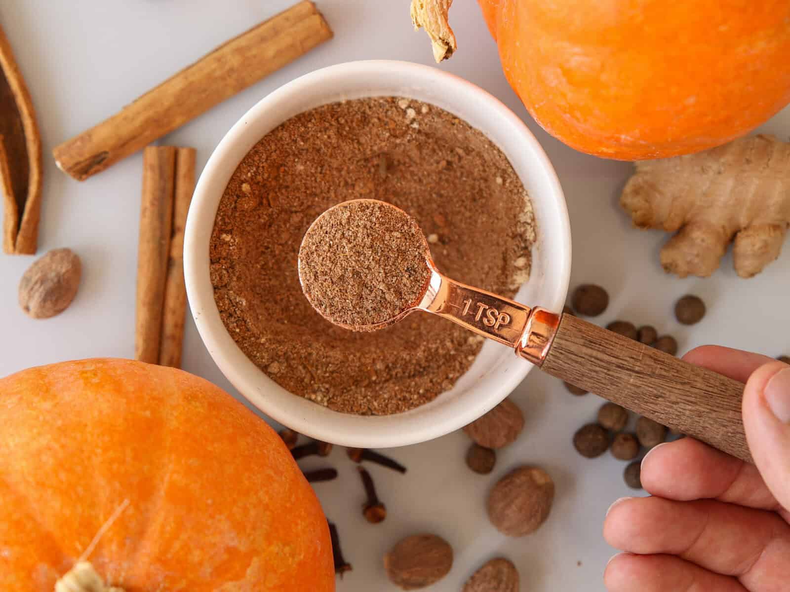 A hand holds a teaspoon of pumpkin spice mix above a small bowl, surrounded by pumpkins, cinnamon sticks, nutmeg, ginger root, and whole cloves on a white surface.