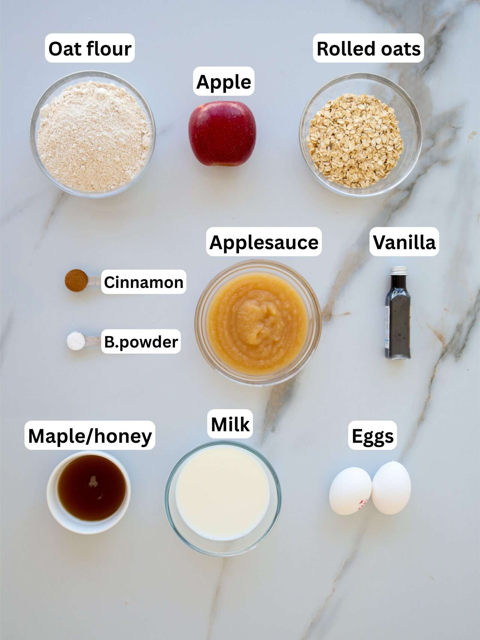 A flat lay of labeled baking ingredients: oat flour, apple, rolled oats, cinnamon, baking powder, applesauce, vanilla, maple syrup or honey, milk, and two eggs, all arranged on a light surface.