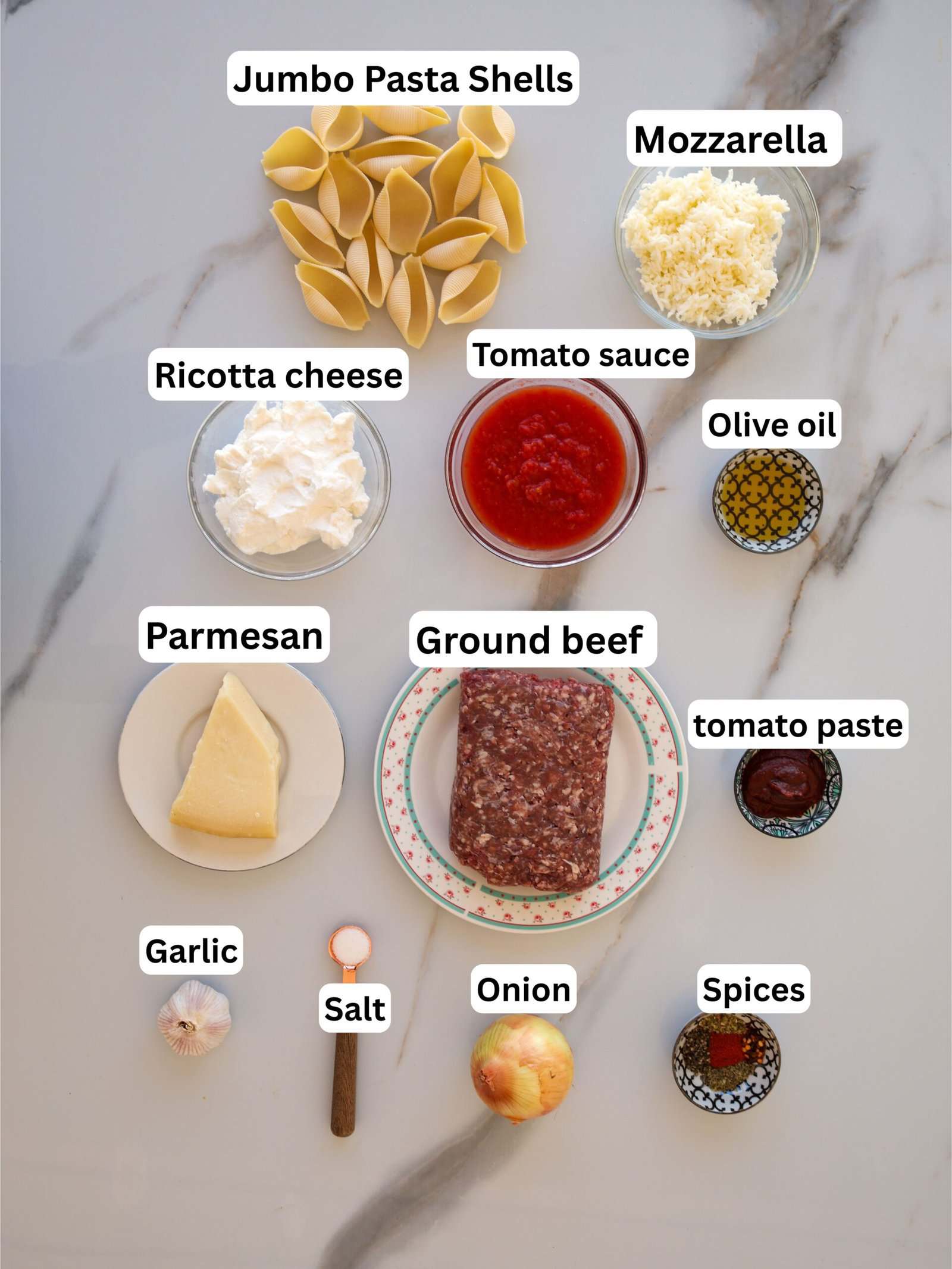 Labeled ingredients for stuffed pasta shells are arranged on a surface: jumbo pasta shells, mozzarella, ricotta cheese, tomato sauce, olive oil, parmesan, ground beef, tomato paste, garlic, salt, onion, and spices.