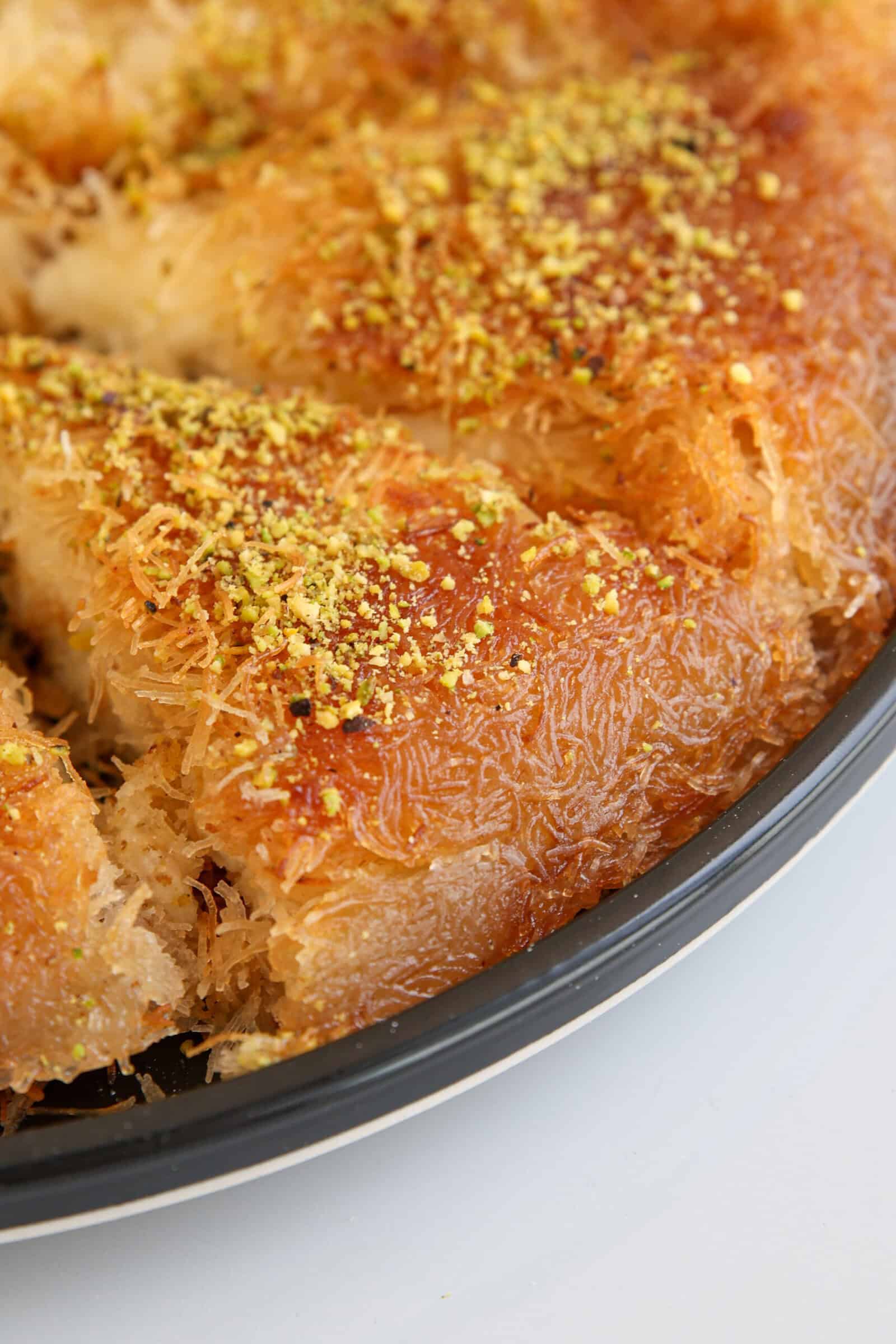 Close-up of a round tray of kunafa, a Middle Eastern dessert made with shredded phyllo dough, golden brown and topped with crushed pistachios. A few slices have been cut and separated.
