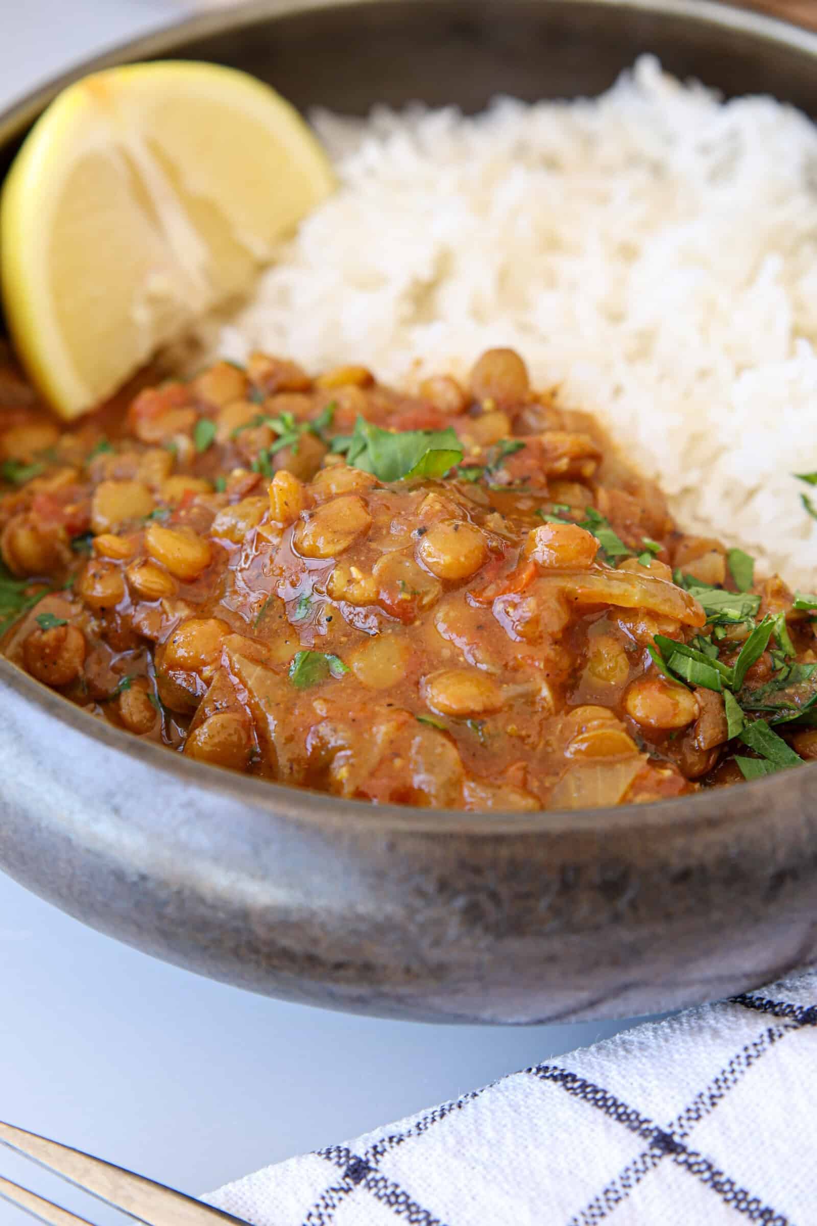 A close-up of a bowl filled with white rice and lentil curry, garnished with chopped herbs and served with a lemon wedge on the side. A checkered napkin is partially visible under the bowl.