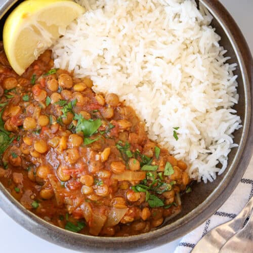 A bowl of white rice and lentil curry garnished with chopped herbs and a lemon wedge, placed next to a striped napkin and metal cutlery.