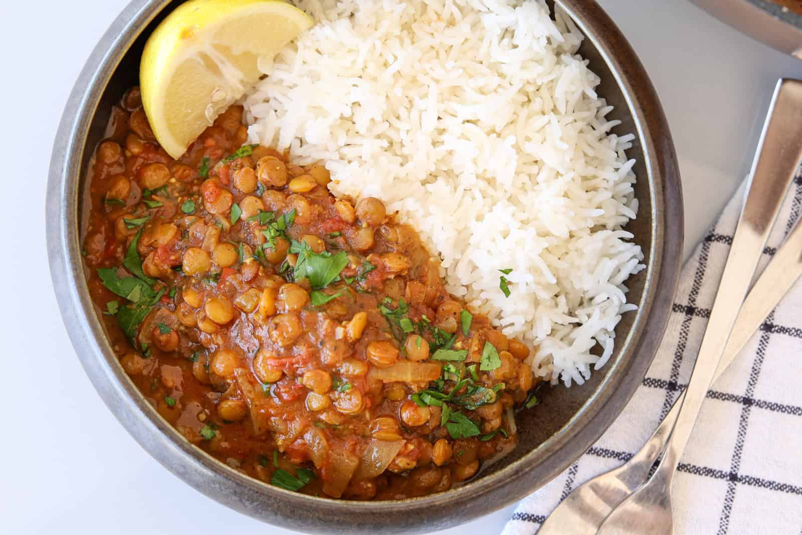 A bowl of white rice and lentil curry garnished with chopped herbs and a lemon wedge, placed next to a striped napkin and metal cutlery.