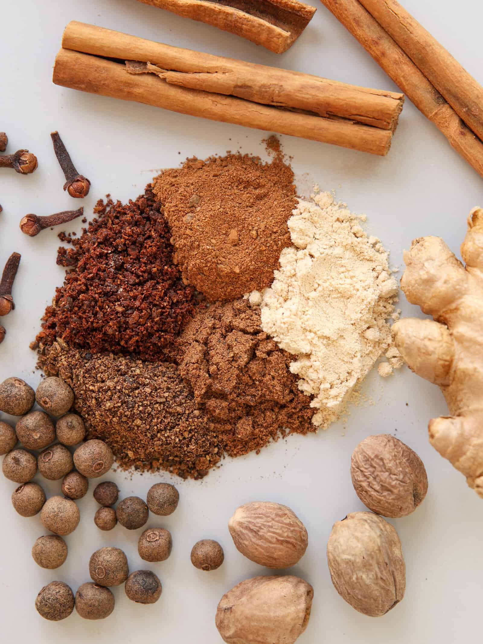 A flat lay of whole and ground spices on a white surface, including cinnamon sticks, cloves, ginger root, allspice, nutmeg, and piles of ground ginger, cinnamon, nutmeg, cloves, and allspice.