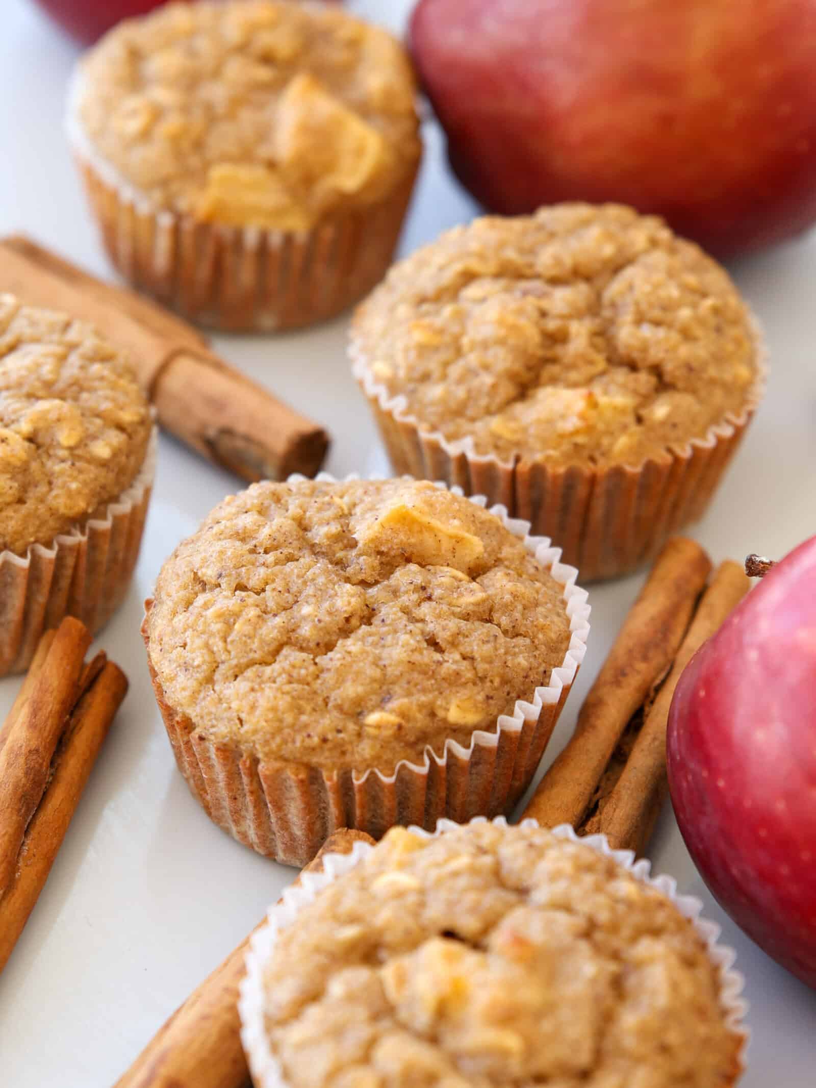 Apple oatmeal muffins in paper liners are arranged on a white surface, surrounded by whole red apples and cinnamon sticks. The muffins have a golden-brown, textured top with visible apple pieces.
