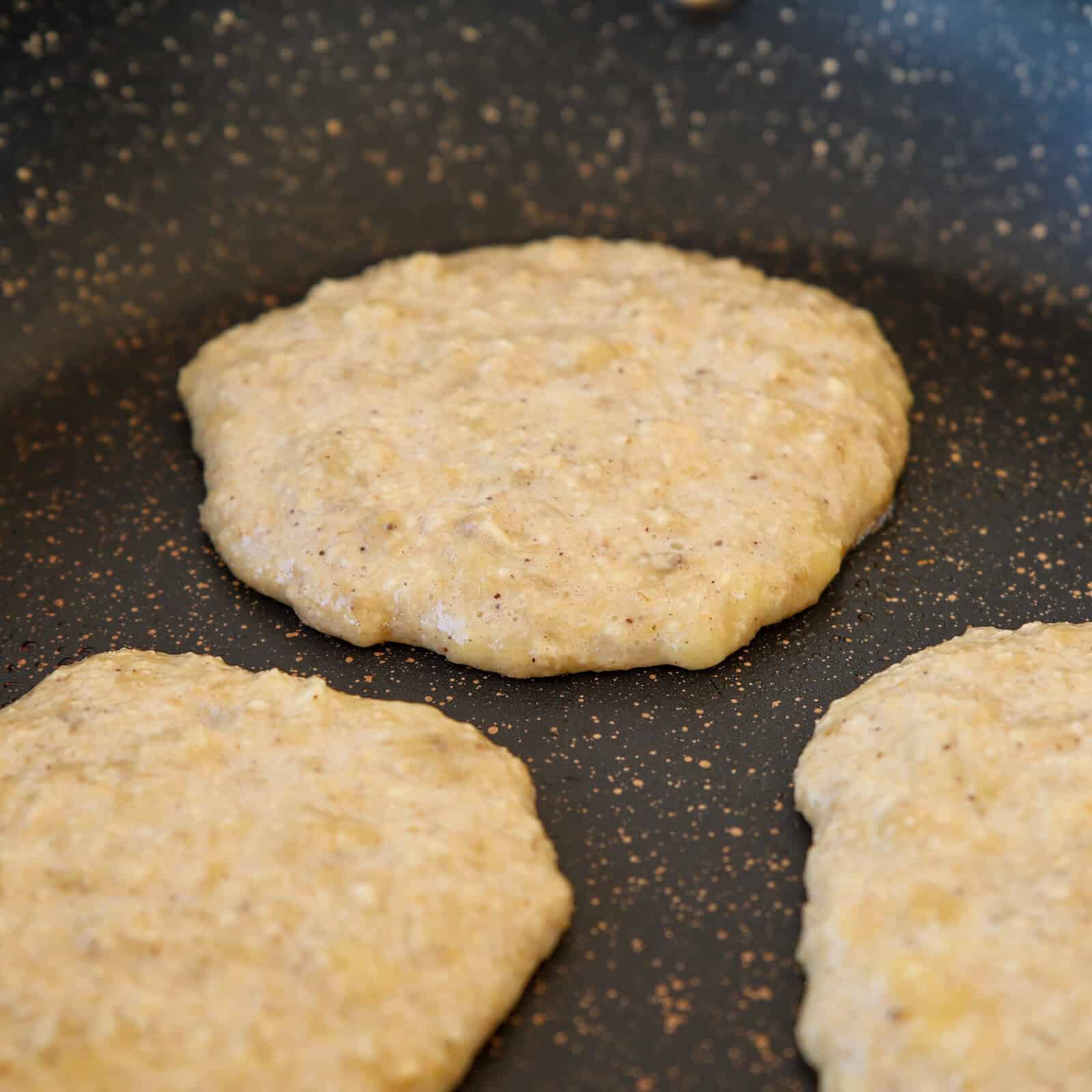 Three uncooked pancakes made from a coarse, grainy batter are cooking on a speckled, nonstick frying pan surface. The pancakes are round and slightly uneven in shape.