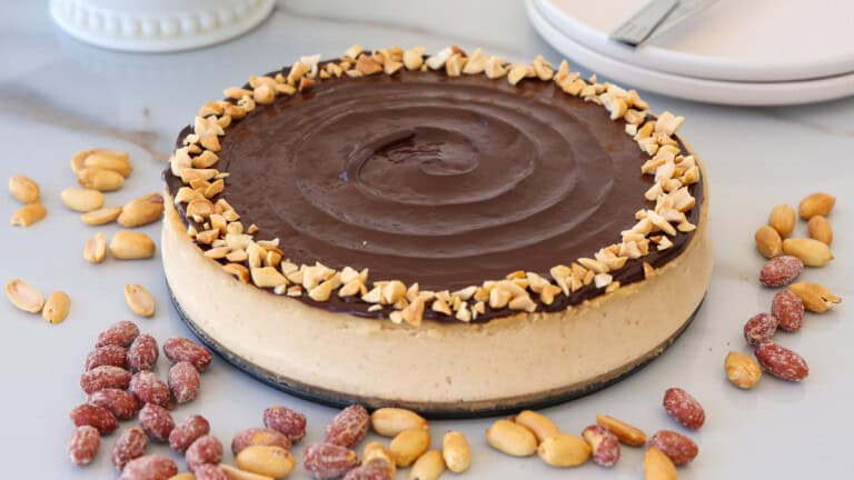 A round peanut butter cheesecake topped with a smooth layer of chocolate ganache and a border of chopped peanuts, surrounded by scattered whole peanuts on a white surface.