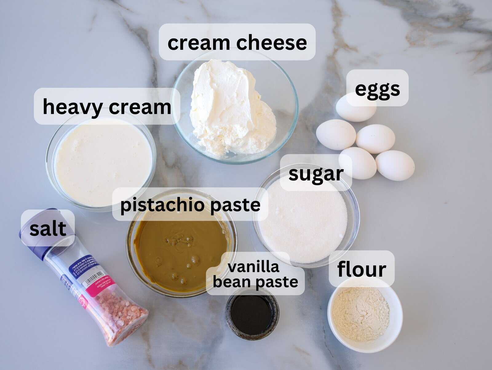 A marble countertop displays labeled ingredients in bowls: cream cheese, eggs, heavy cream, sugar, flour, pistachio paste, vanilla bean paste, and a container of salt.