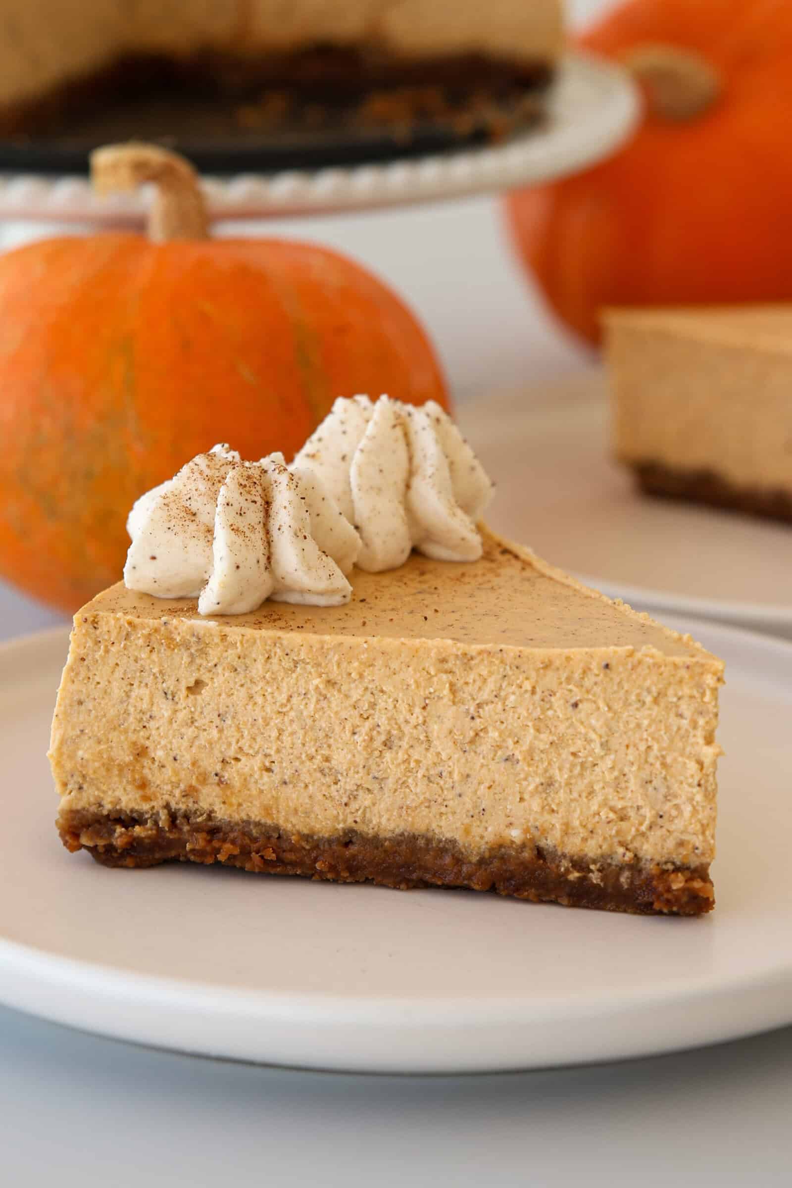 A slice of pumpkin cheesecake with a graham cracker crust, topped with whipped cream and a sprinkle of cinnamon, sits on a white plate. Two small pumpkins are in the background.