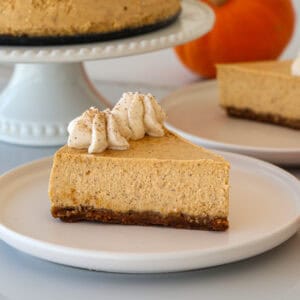 A slice of pumpkin cheesecake with a graham cracker crust, topped with whipped cream and a sprinkle of cinnamon, sits on a white plate; a whole cheesecake and pumpkins are in the background.