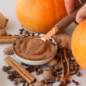 A hand holds a spoon of pumpkin pie spice above a bowl, surrounded by whole spices like cinnamon sticks, cloves, nutmeg, allspice, ginger, and pumpkins, suggesting a fall or pumpkin spice blend.
