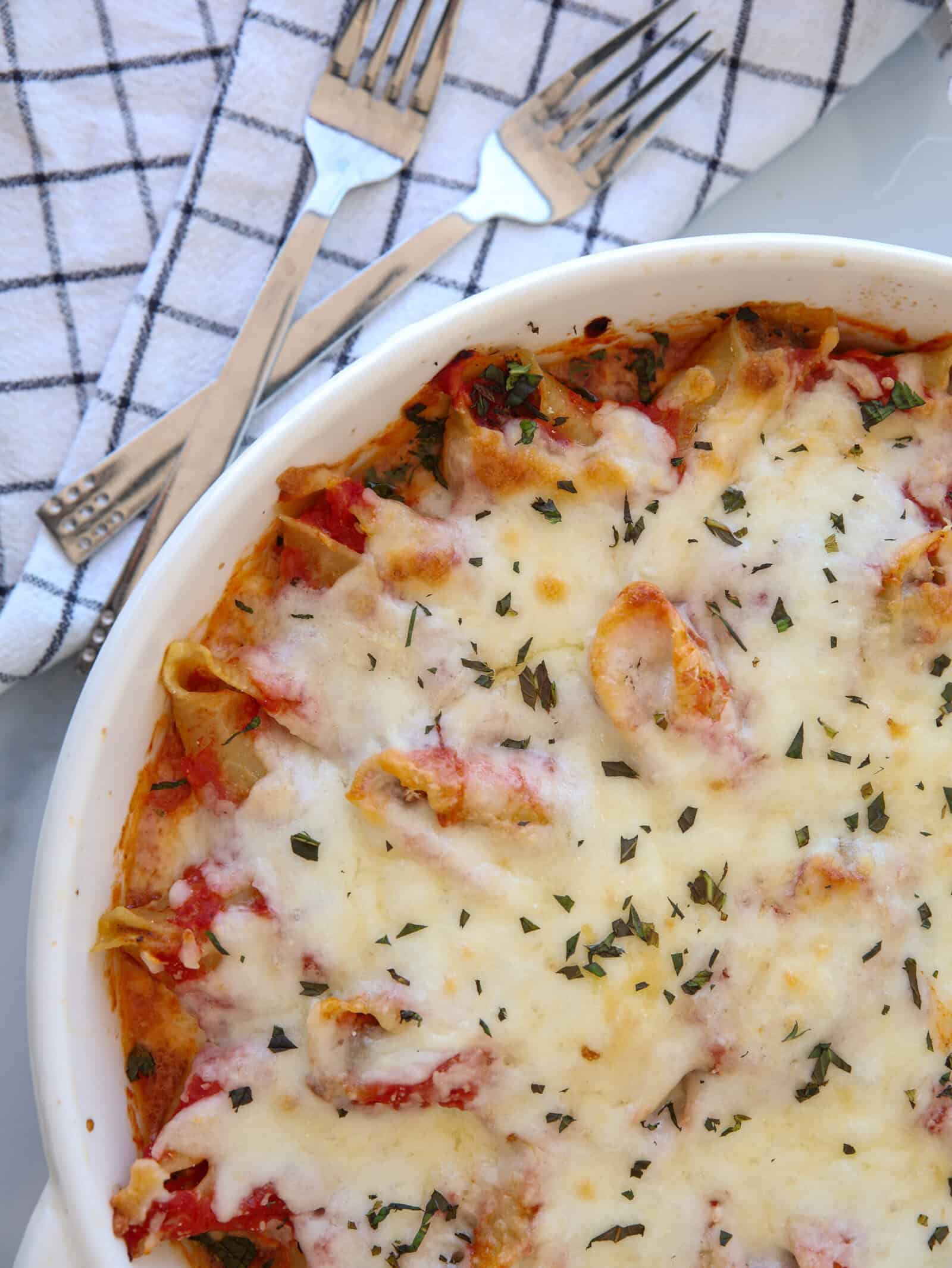 A baked pasta dish topped with melted cheese and herbs sits in a white round casserole dish. Three forks rest on a checkered cloth nearby.