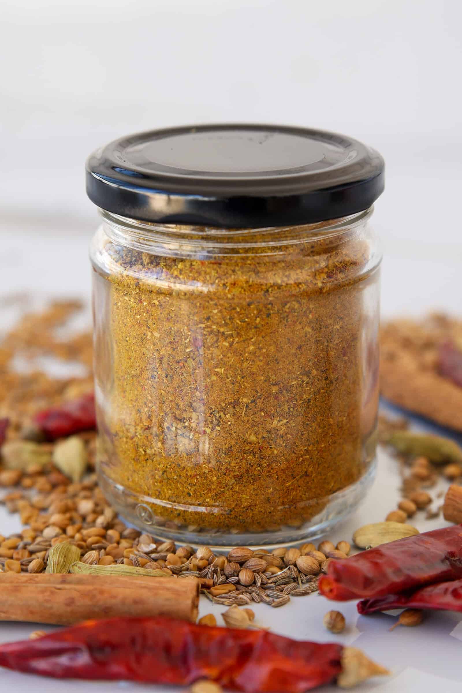 A glass jar filled with golden-brown curry powder stands on a surface scattered with dried red chilies, coriander seeds, cardamom pods, cinnamon sticks, and other whole spices. The jar has a black lid.