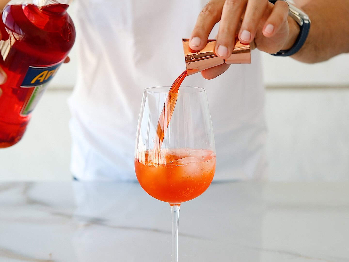 A person in a white shirt pours a red-orange liquid from a jigger into a wine glass filled with ice on a marble countertop. A bottle labeled "Aperol" is visible in their other hand.