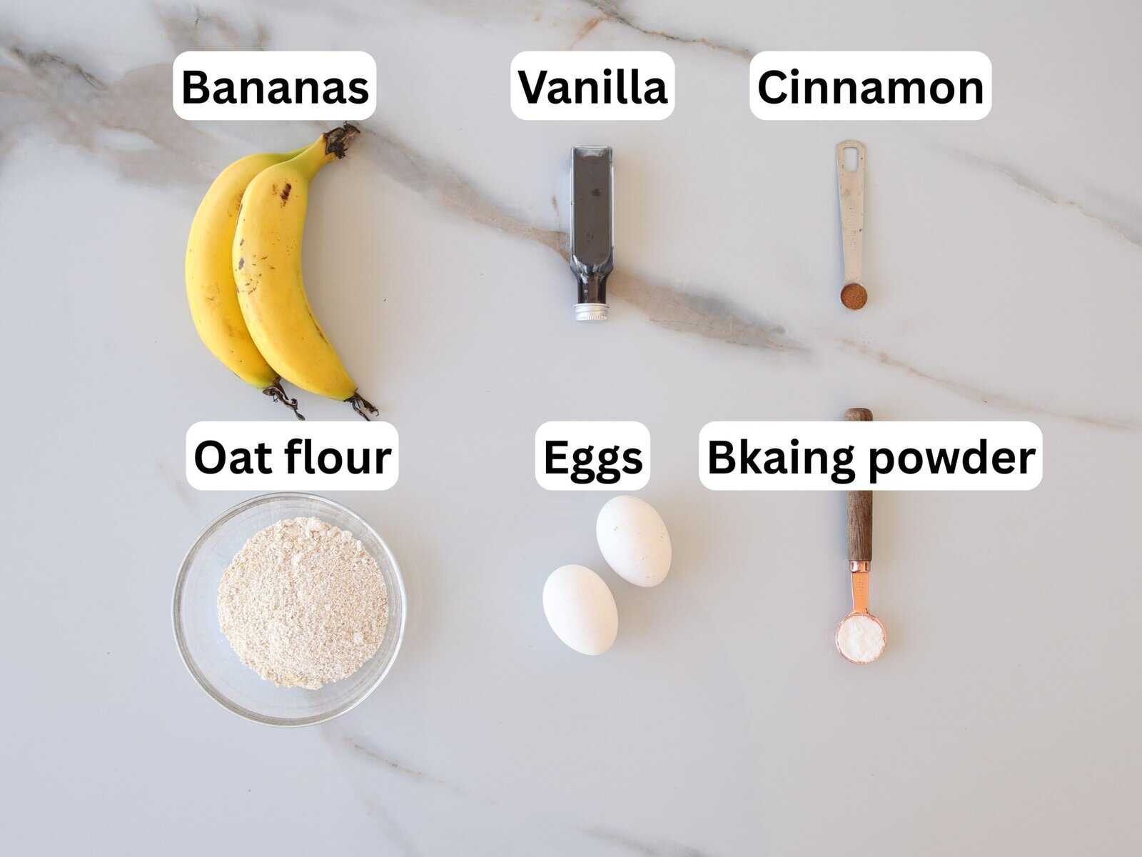 Six labeled baking ingredients on a marble counter: three bananas, a tube of vanilla, a teaspoon of cinnamon, a bowl of oat flour, two eggs, and a measuring spoon of baking powder.