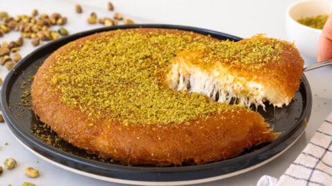 A round kunafa dessert topped with ground pistachios on a black plate. A slice is being lifted, revealing the gooey cheese filling inside. Pistachios and a bowl of more ground pistachios are in the background.