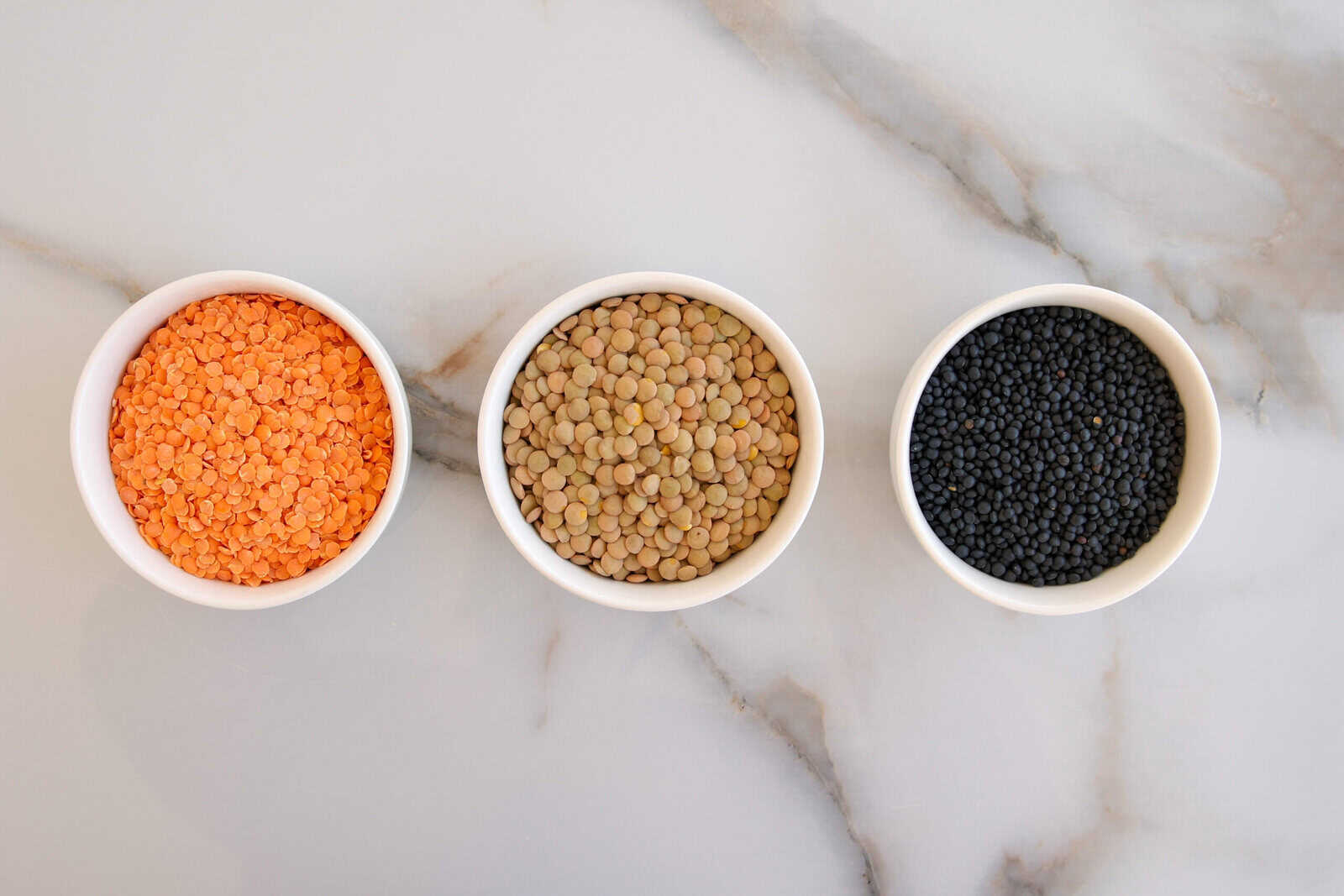 Three white bowls filled with different types of lentils—red, brown, and black—are arranged in a row on a light marble surface.