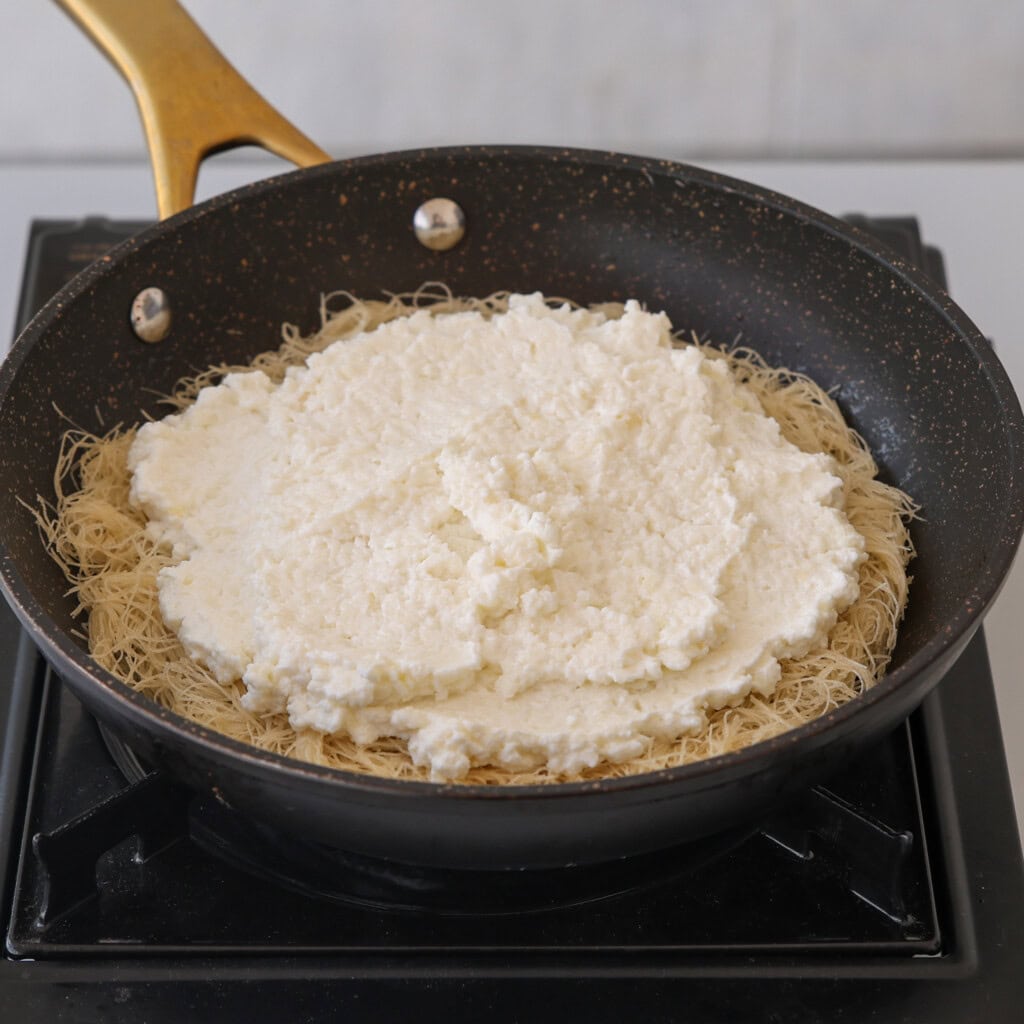 A nonstick frying pan on a stovetop contains a layer of shredded pastry topped with a thick layer of white cheese mixture, ready to be cooked.