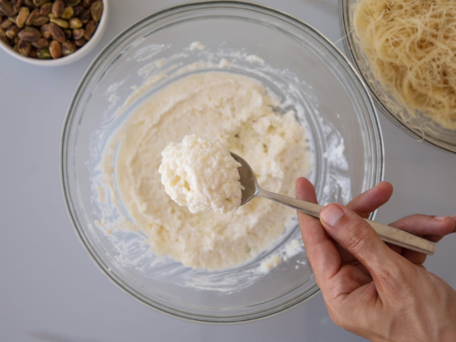 A hand holds a spoonful of creamy ricotta cheese mixture above a glass bowl, with shredded pastry and a bowl of pistachios nearby on a light surface.