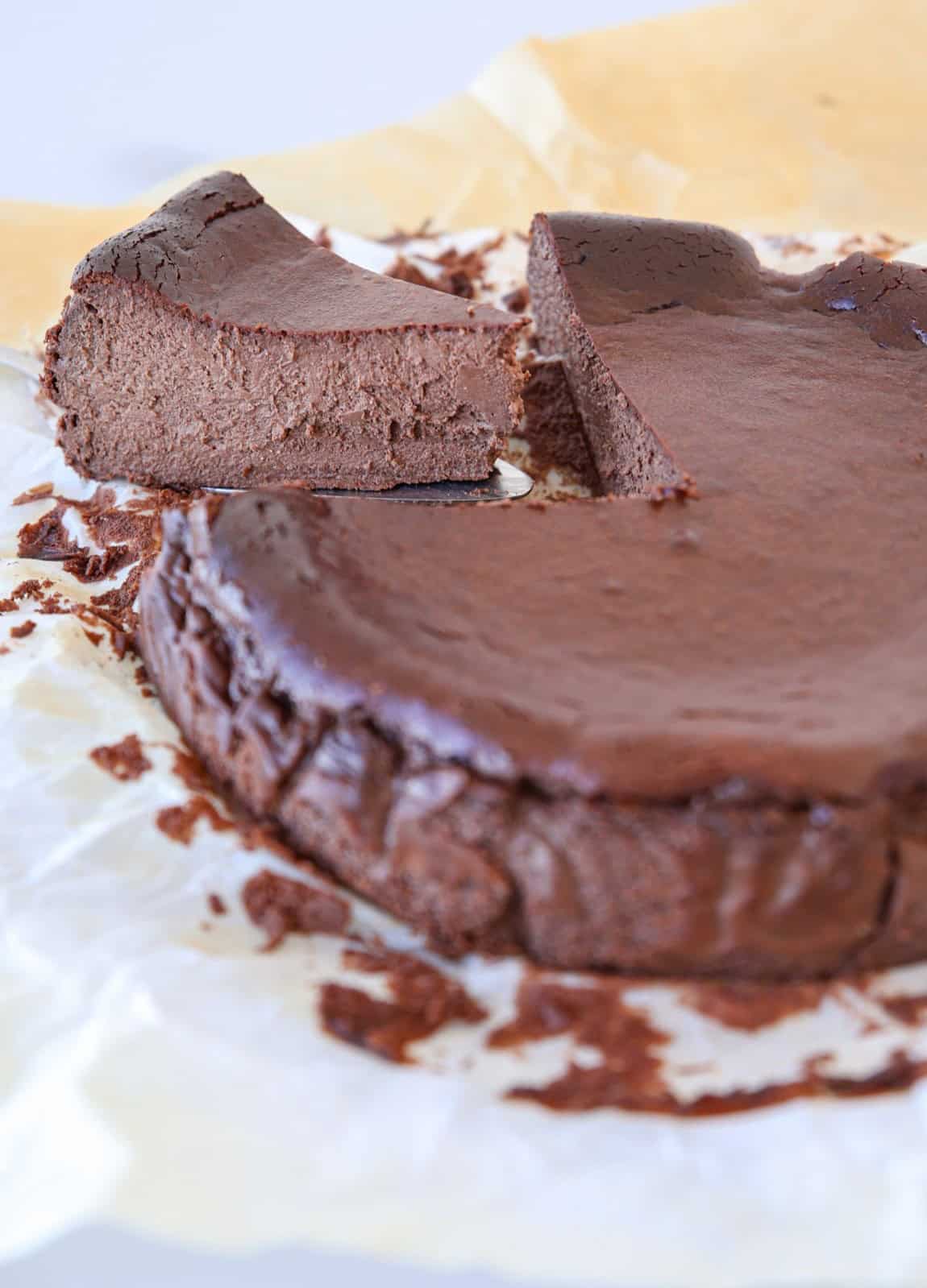 A rich, creamy dark chocolate Basque cheesecake with a dense, fudgy texture sits on parchment paper. A large slice has been cut and slightly lifted, revealing the moist interior of the cake.
