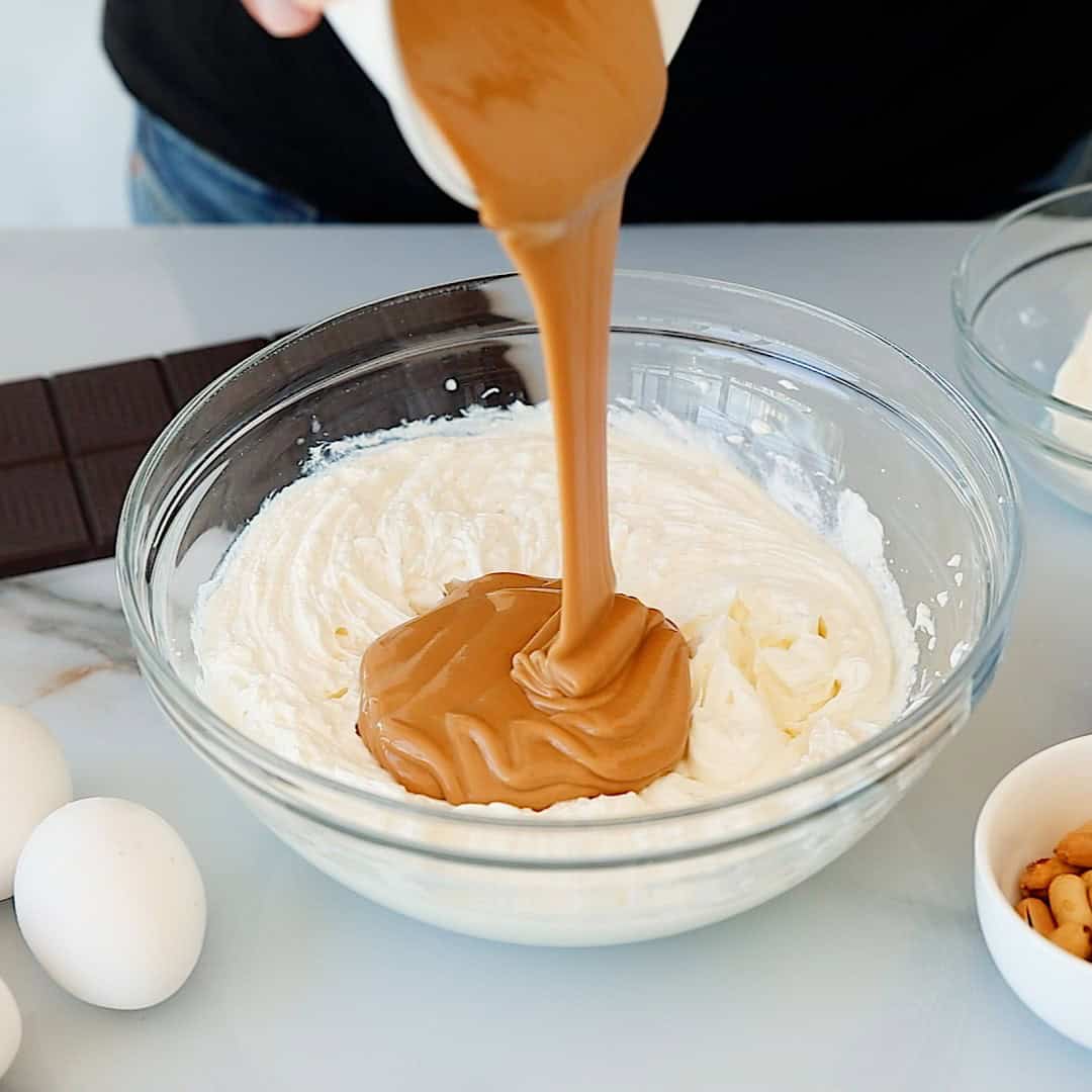 A person pours melted chocolate into a glass bowl filled with creamy white batter, surrounded by eggs, chocolate, and a small bowl of nuts on a white countertop.