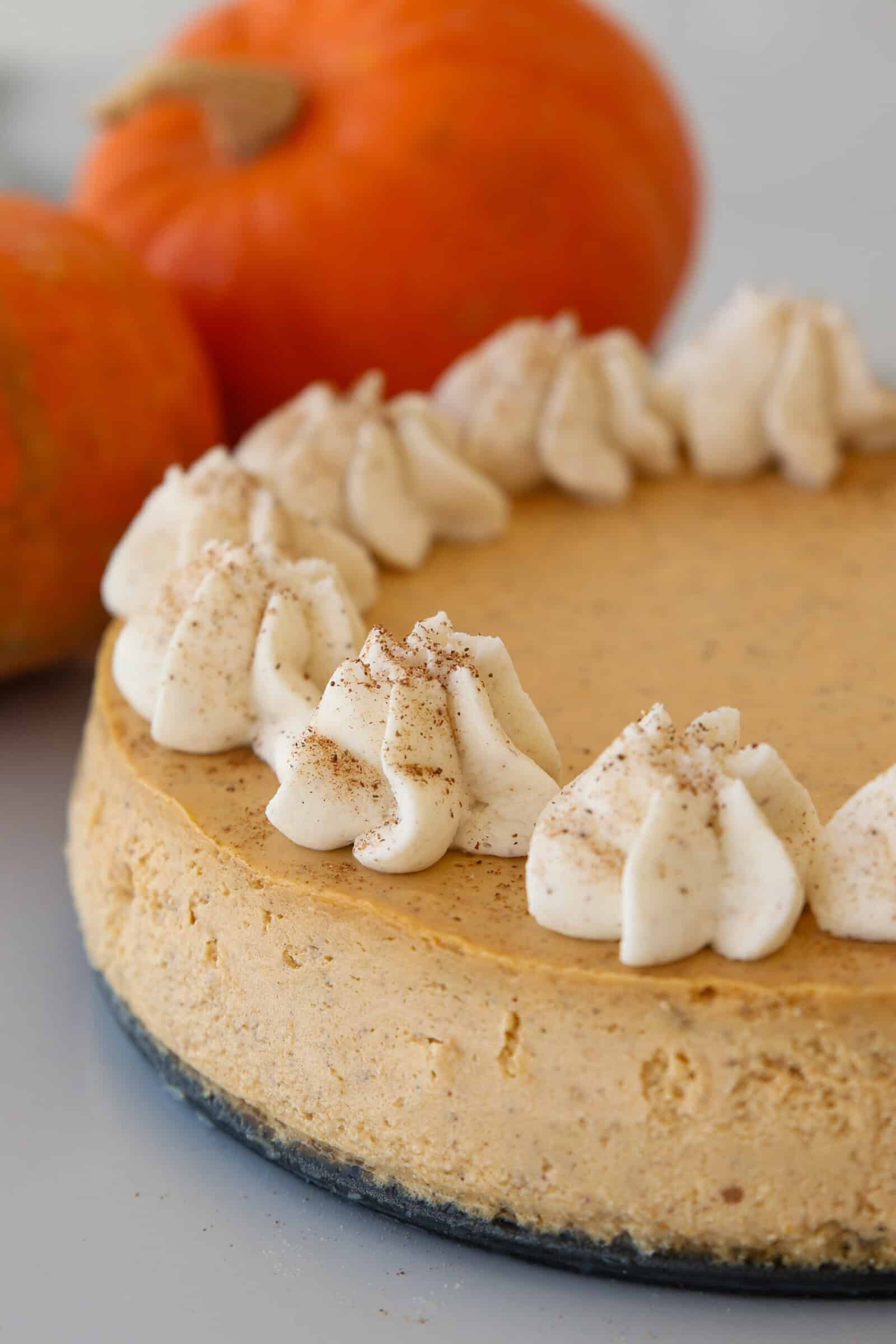 A baked pumpkin cheesecake topped with swirls of whipped cream and a sprinkle of spice, with two pumpkins in the background.
