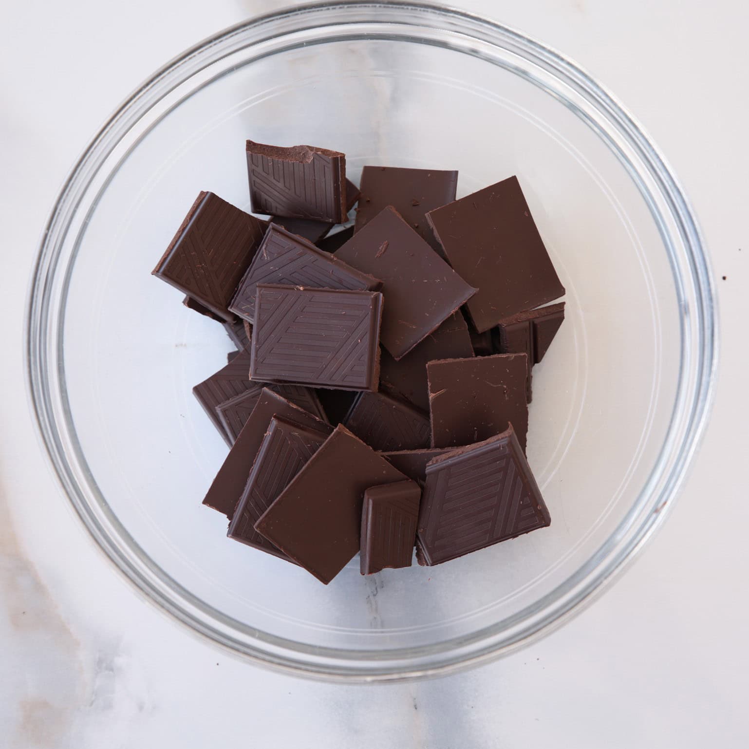 A clear glass bowl filled with several pieces of dark chocolate, each cut into square shapes, sits on a white marble surface.