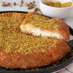 A round, golden-brown knafeh dessert topped with crushed pistachios, with one slice cut and slightly pulled out, revealing a creamy white filling. A bowl of extra crushed pistachios is in the background.
