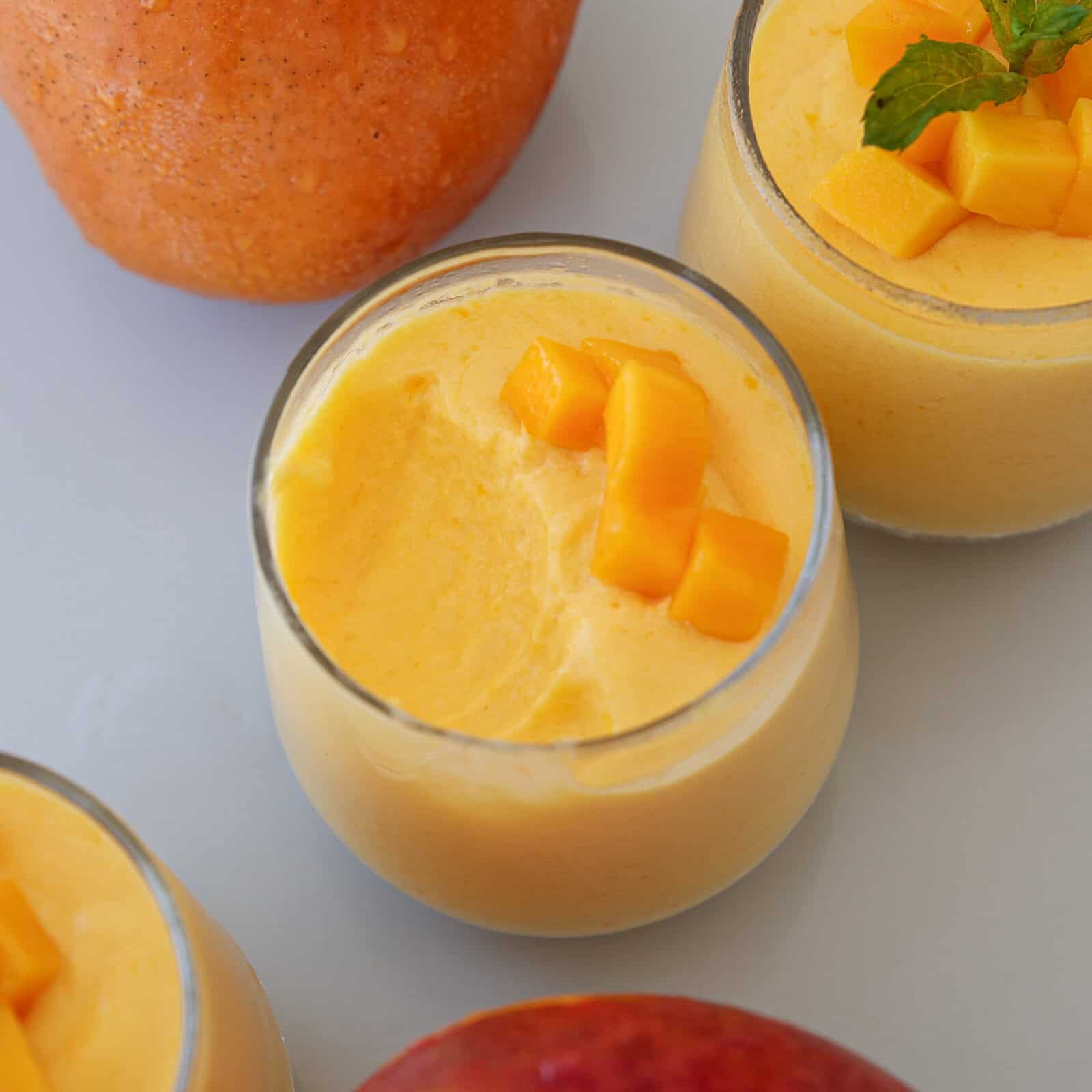Top view of glasses filled with creamy mango mousse, garnished with diced mango and mint. Whole mangoes are placed nearby on a light surface.