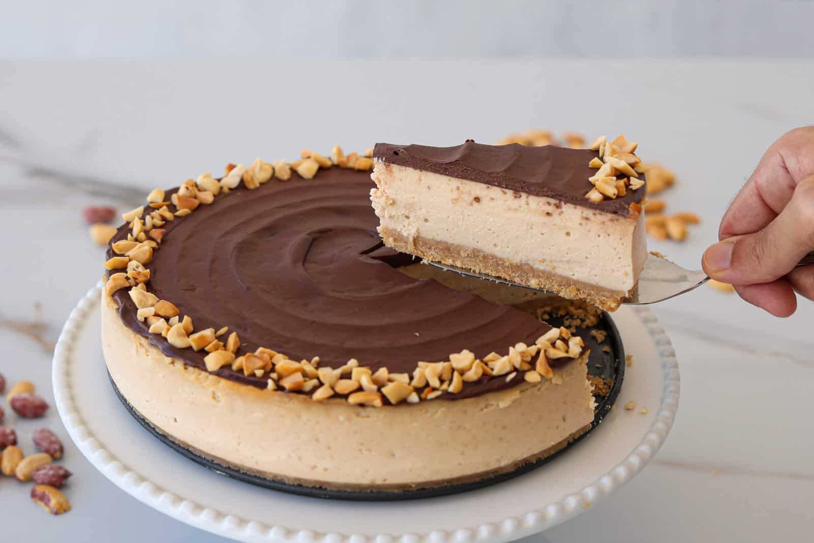 A hand lifts a slice of a creamy cheesecake topped with chocolate and chopped peanuts from a whole cheesecake on a white cake stand. Some chopped peanuts are scattered on the surface nearby.