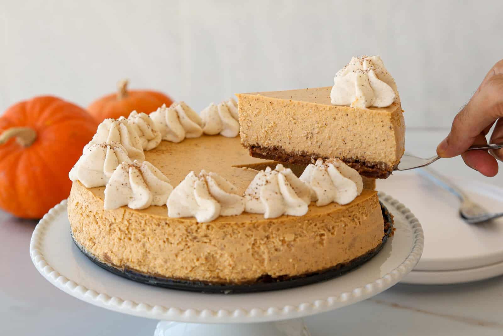 A pumpkin pie cheesecake topped with dollops of whipped cream sits on a cake stand. A hand lifts a slice, and two small pumpkins are visible in the background.